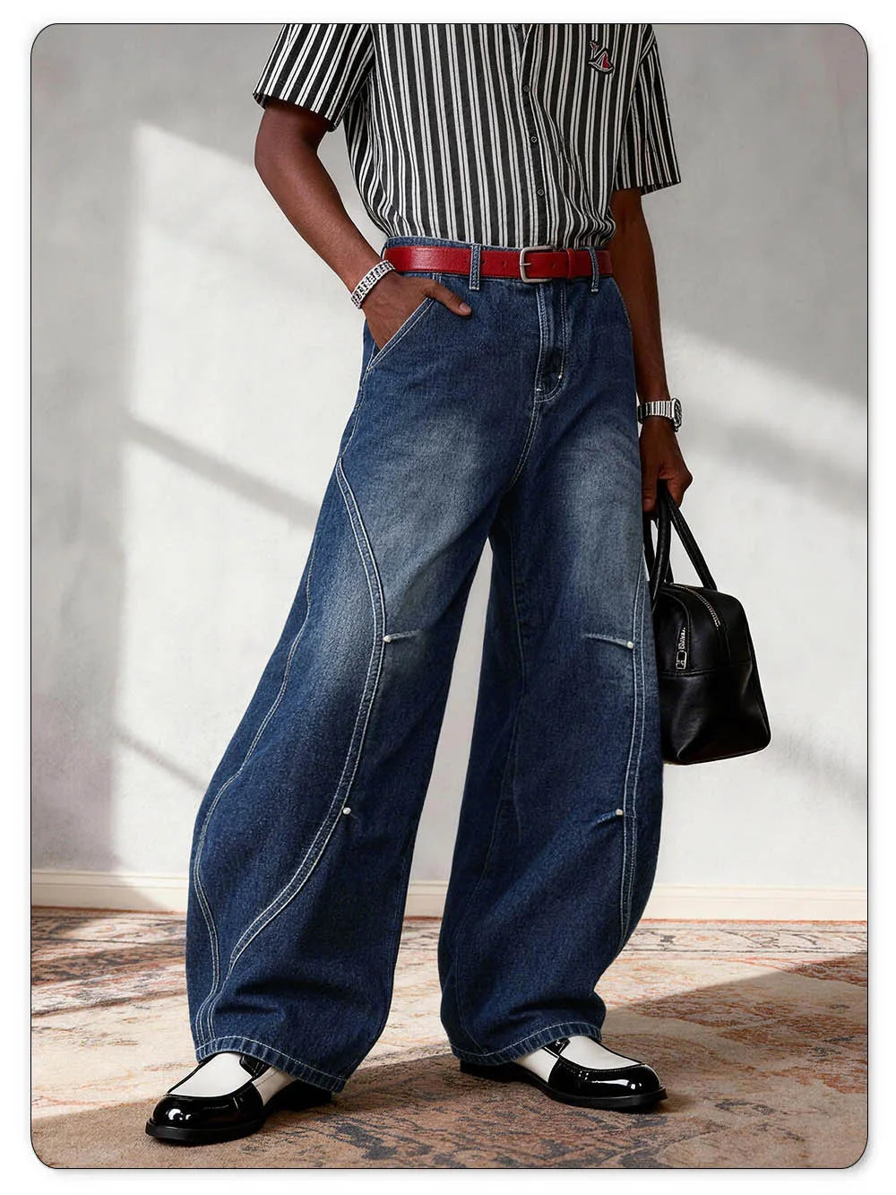 A person wearing oversized dark blue denim jeans with diagonal seam details, styled with a red belt, a black-and-white striped short-sleeve shirt, black shoes, and holding a black handbag.