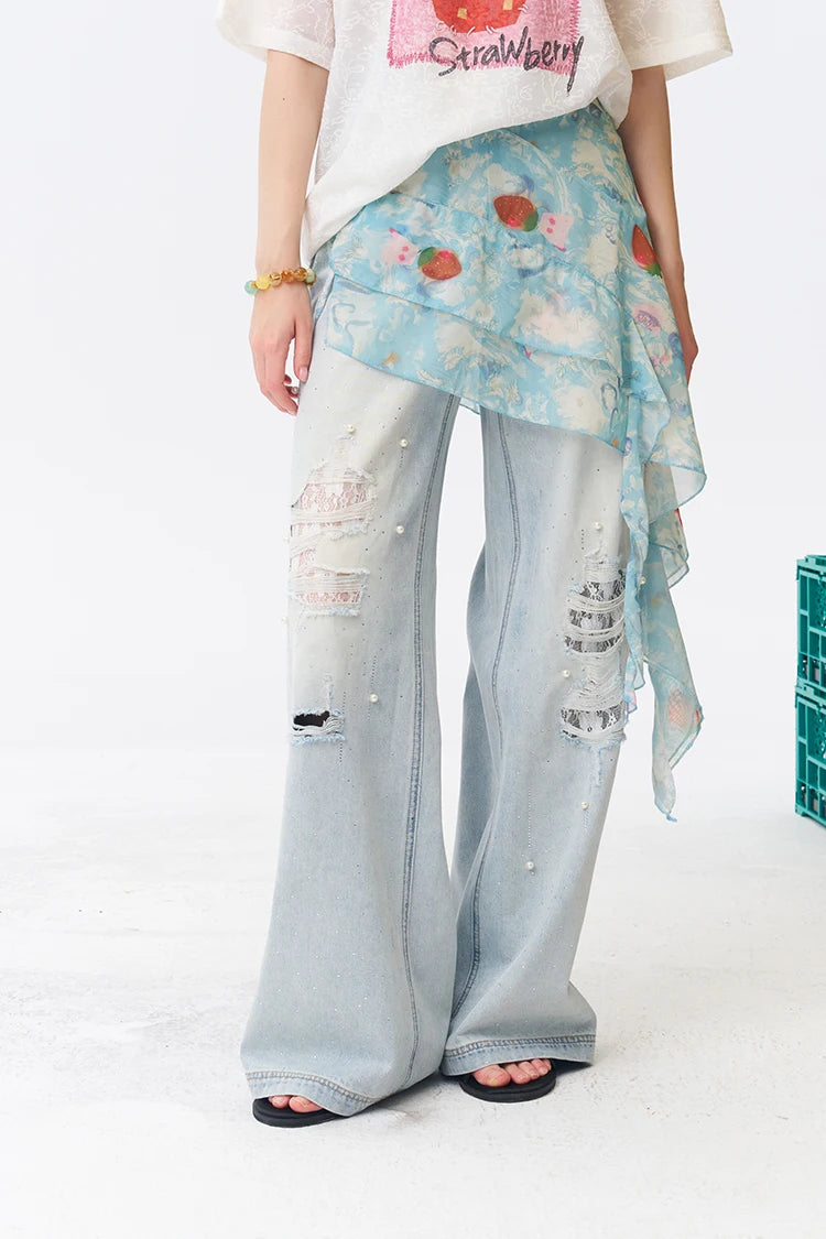 A person wearing light-wash distressed wide-leg jeans with a sheer light blue floral wrap draped over the hips, paired with a white graphic T-shirt and black sandals, photographed against a clean white studio background.