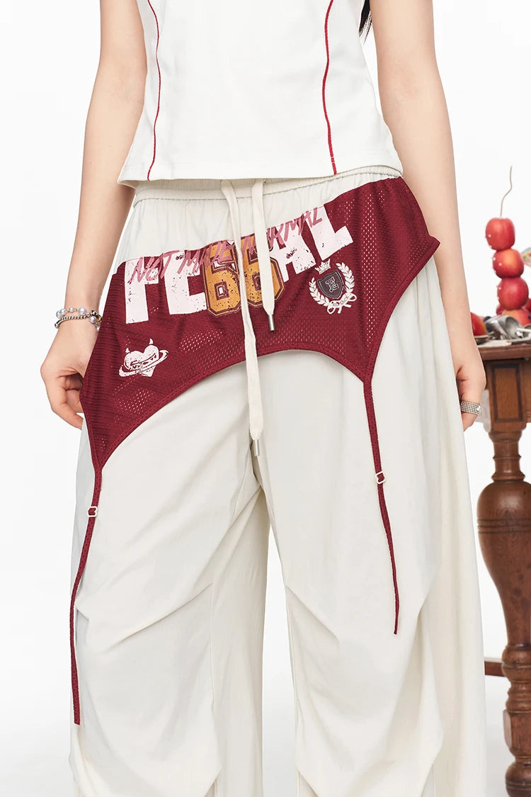 A person wearing loose off-white wide-leg pants with a large maroon mesh side panel, white drawstring waist, and bold white lettering across the front, paired with a white sleeveless top.
