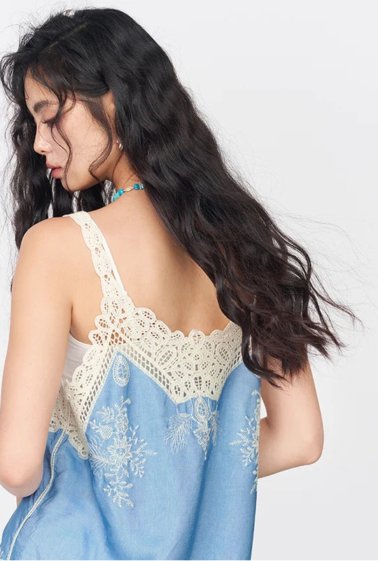 A woman shown from the back wearing a light blue camisole-style top with cream lace trim and floral embroidery, against a plain light gray studio background.
