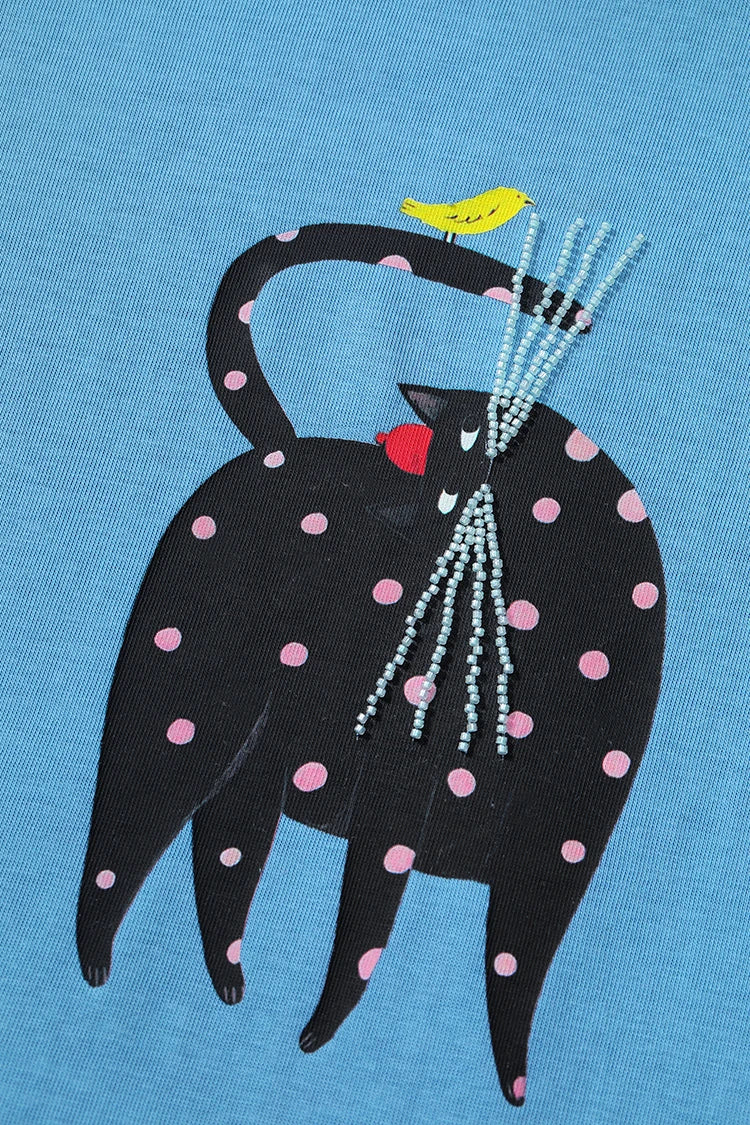 A close-up of a light blue fabric featuring a graphic of a black cat with pink polka dots, a small yellow bird on its back, and a bundle of silver beaded whiskers.