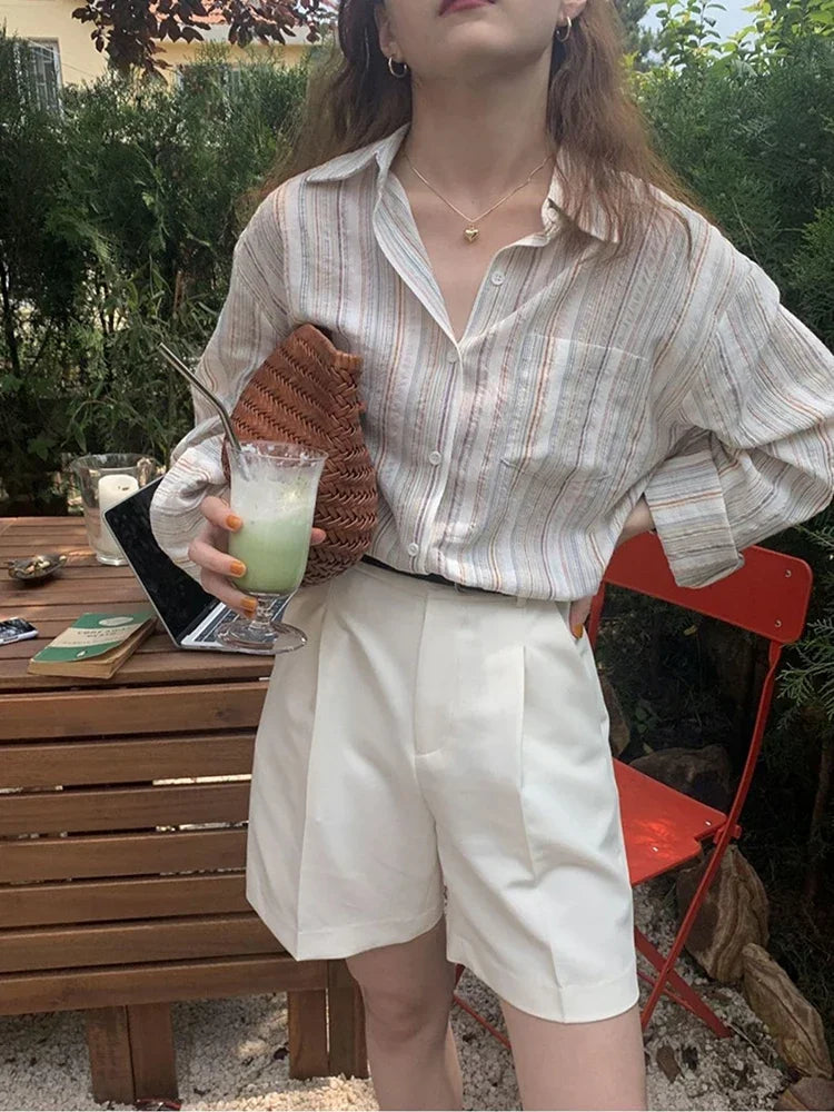 Person holding a drink outdoors, wearing a striped shirt and white shorts.