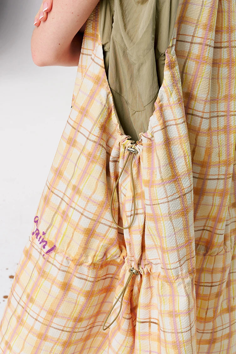 A close-up of a plaid fabric garment, likely a dress or long wrap, in soft yellow, peach, and white tones, with a drawstring closure and a taupe inner layer visible at the top. A person's arm is visible on the left side.