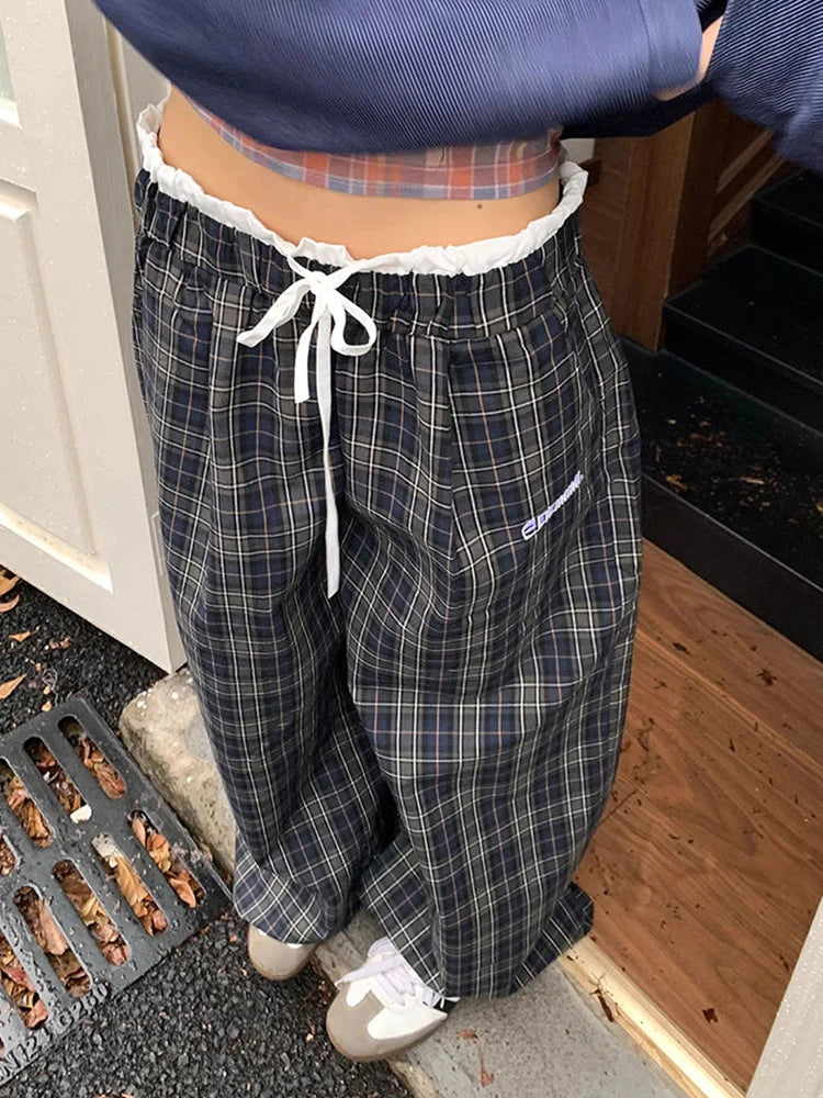 A person wearing loose, wide-leg plaid pants in dark navy and gray with thin white stripes, featuring a white drawstring waistband and white sneakers.