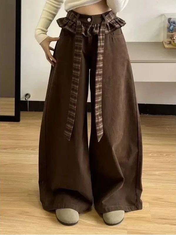 A person wearing wide-leg brown pants with a plaid waist tie belt, styled with a cream ribbed crop top and gray slip-on shoes in an indoor room with light wood flooring.