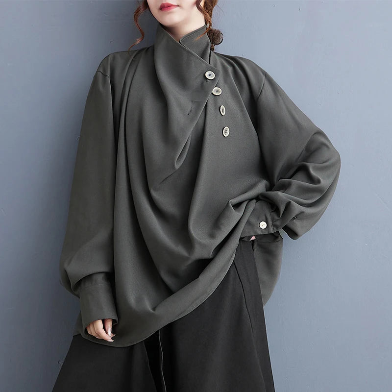 A woman wearing a dark olive green high-neck blouse with a front button placket and black pants, posing against a textured gray wall.