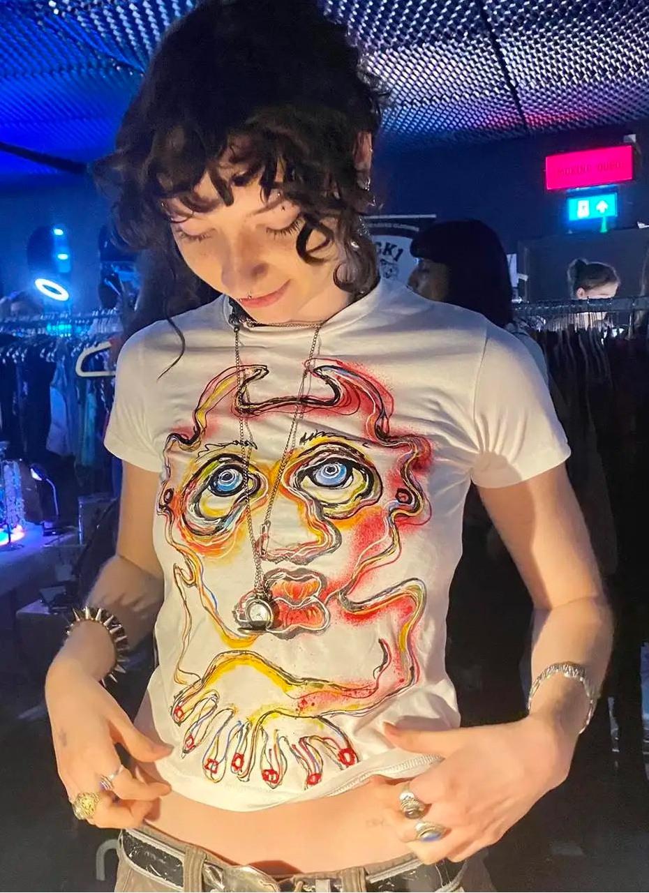 A person wearing a white short-sleeve graphic T-shirt with a large surreal face illustration in red, orange, yellow, and blue, photographed in a dimly lit retail or backstage environment with blue lighting.