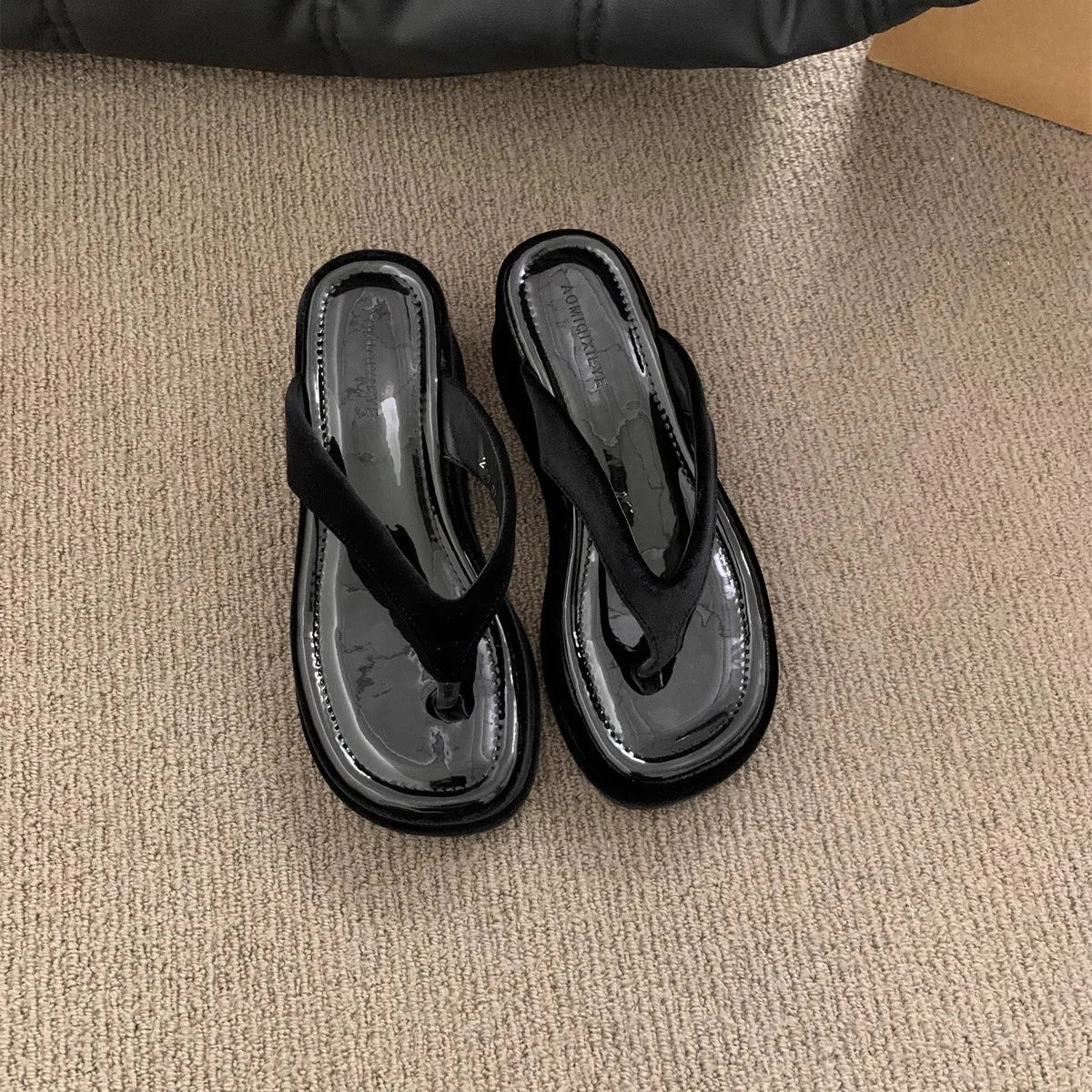 A pair of black patent leather thong sandals with chunky platform soles, resting on a beige carpet.