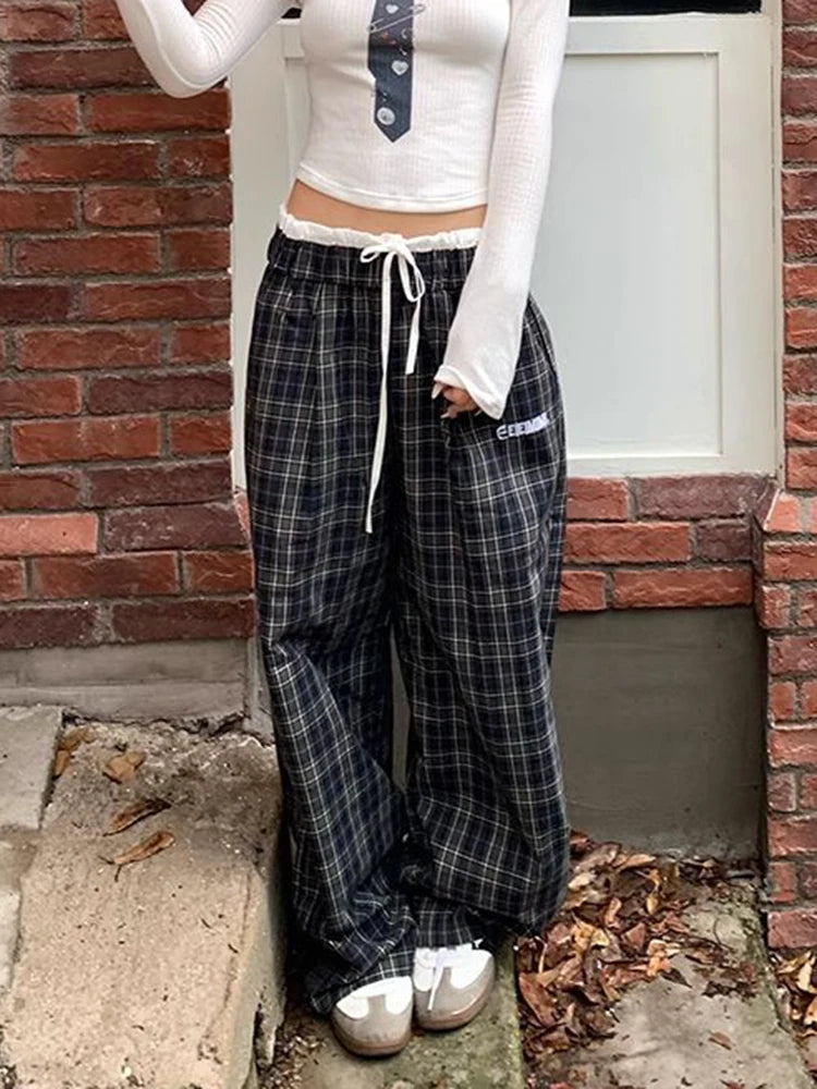 A person wearing black and gray plaid wide-leg sweatpants with a white drawstring, styled with a white long-sleeve crop top and white sneakers, standing outdoors against a brick wall and white door.