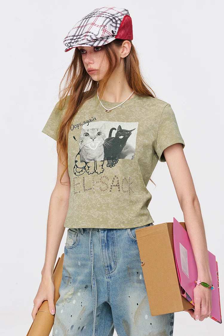 A woman modeling a cropped olive green graphic t-shirt with a cat print, paired with distressed blue jeans, a red and white plaid flat cap, and holding cardboard boxes.