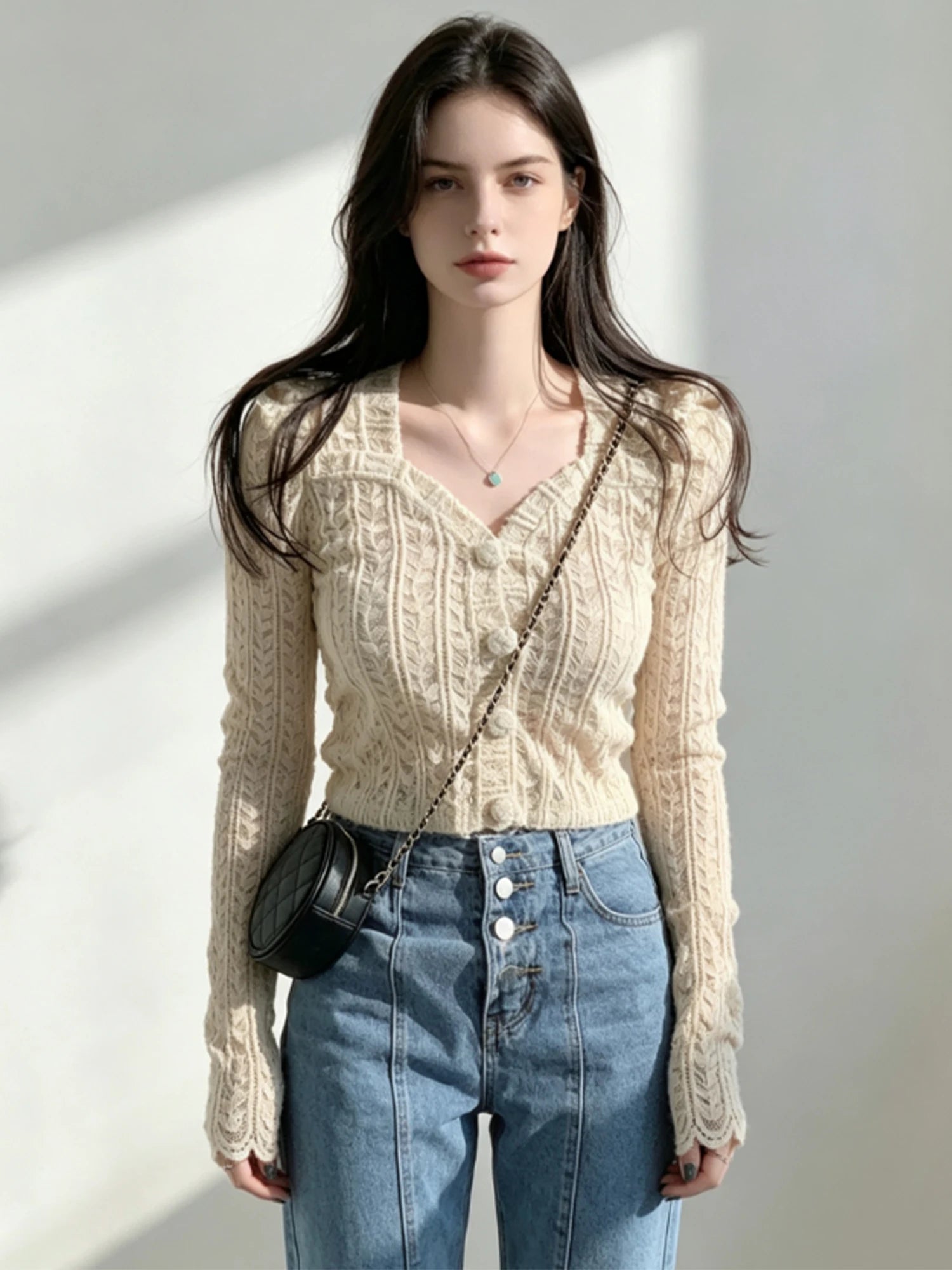 A woman modeling a cream cable-knit V-neck cardigan with a button front, paired with blue jeans and a small black crossbody bag, set against a bright, sunlit neutral wall.