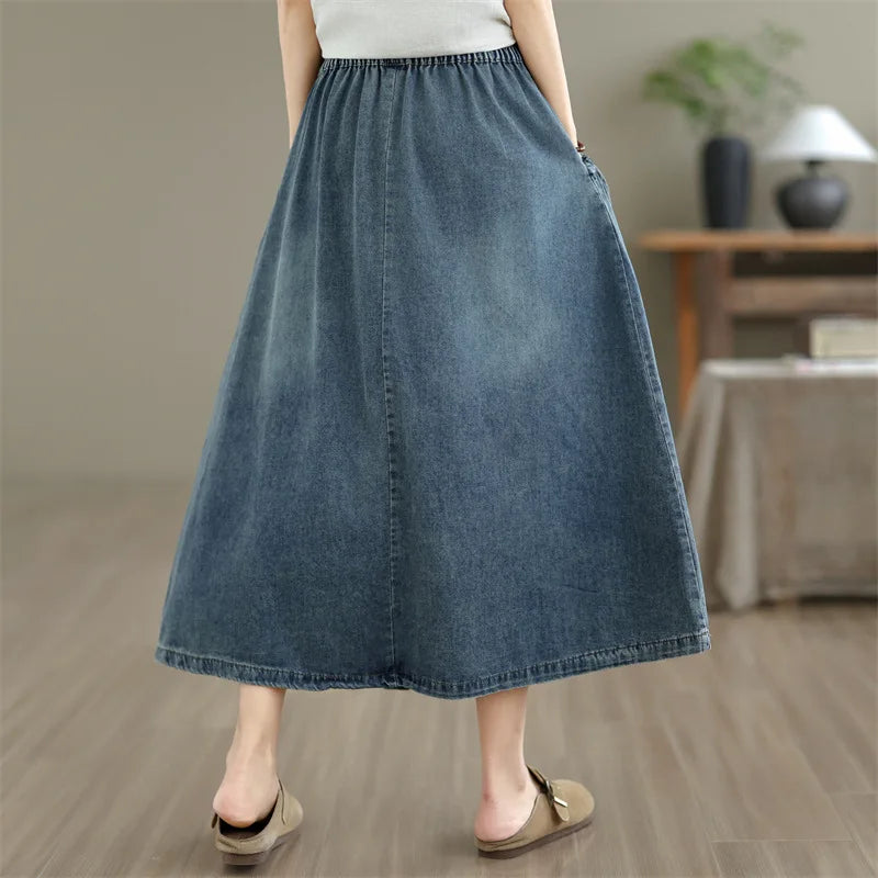 A person wearing a loose, ankle-length blue denim skirt with side pockets, paired with a white top and tan flat sandals, standing in a softly lit indoor room with wood flooring and a table in the background.