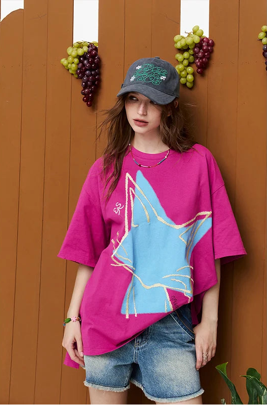 A woman wearing a bright pink oversized T-shirt with a large light blue star graphic outlined in gold, paired with denim shorts and a dark baseball cap, standing against a brown wooden wall with grape decorations.