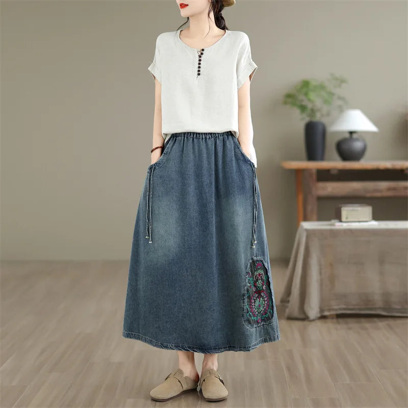 A woman wearing a white short-sleeve top with a buttoned neckline and a high-waisted blue denim maxi skirt featuring side pockets and a colorful embroidered floral detail near the hem, standing in a softly lit indoor room.