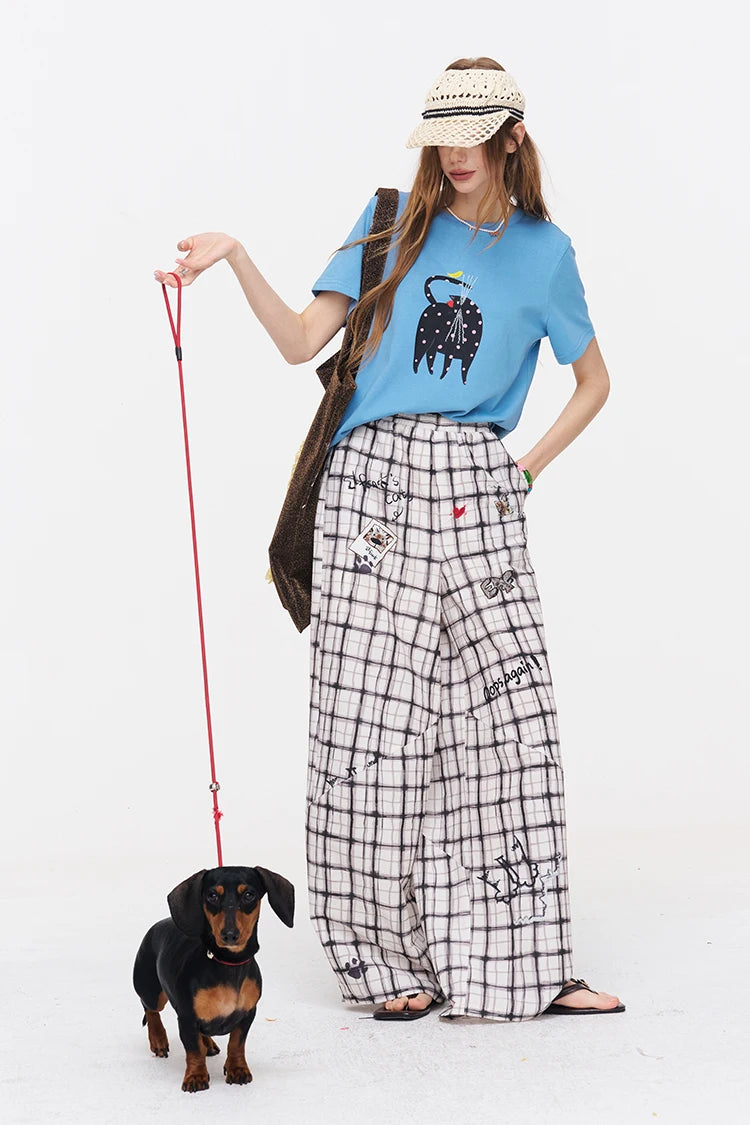 A woman modeling a light blue graphic t-shirt, black-and-white plaid wide-leg pants, and a straw visor, standing next to a small black and tan dachshund on a red leash against a plain white background.