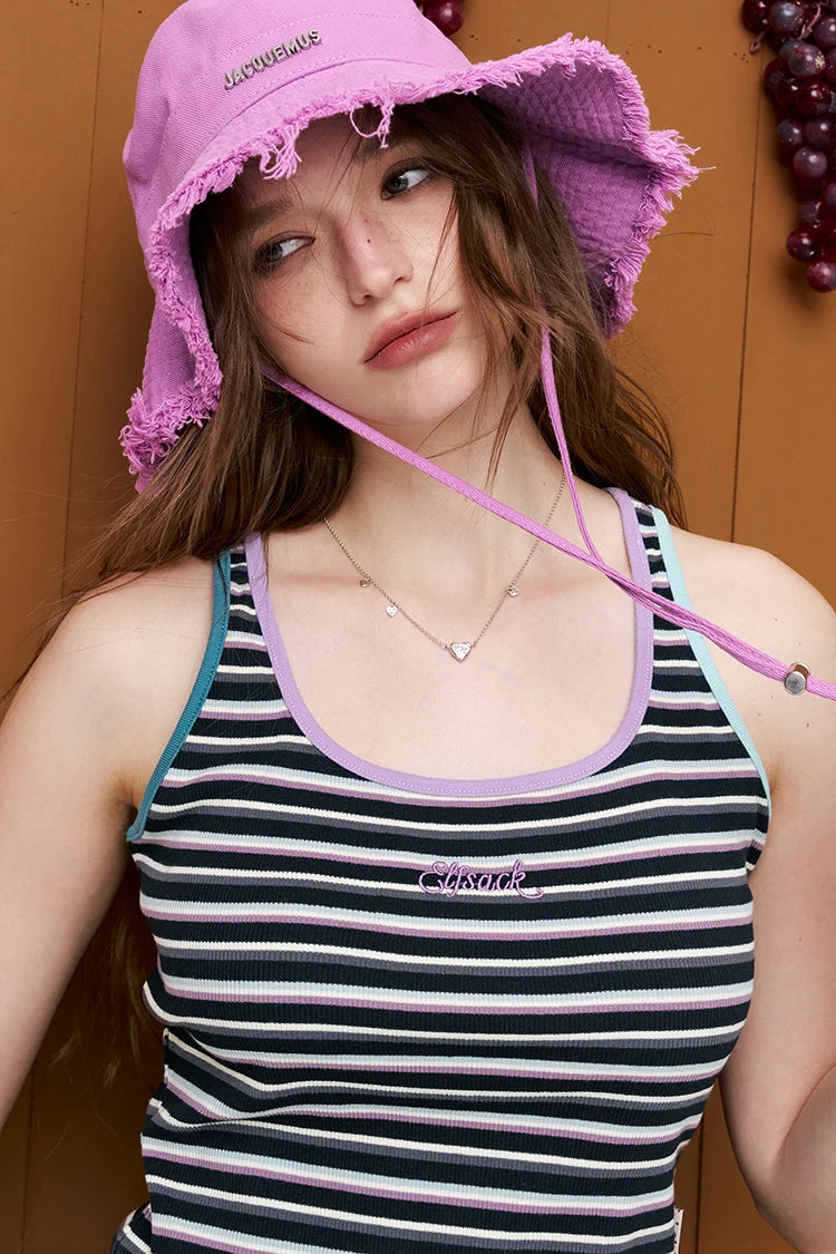 A woman wearing a pink frayed-brim bucket hat with a chin strap and a black, white, and pink striped ribbed tank top, posed against a warm brown wooden background.