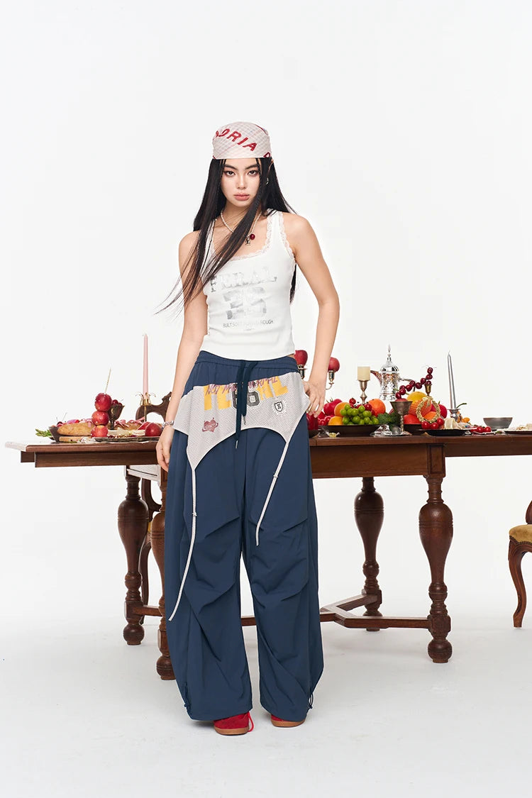 A woman stands in a studio wearing a white graphic tank top, navy wide-leg pants with a white waist panel and drawstring, a white cap with red lettering, and red shoes. A wooden dining table with fruit and candles is behind her against a white background.