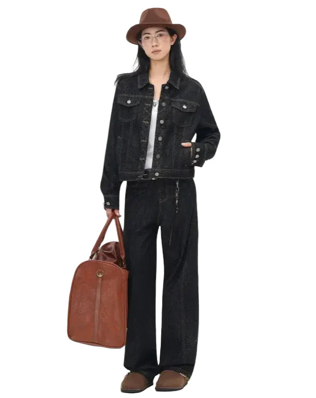 Person wearing a black denim jacket and pants, holding a brown leather bag on a white background
