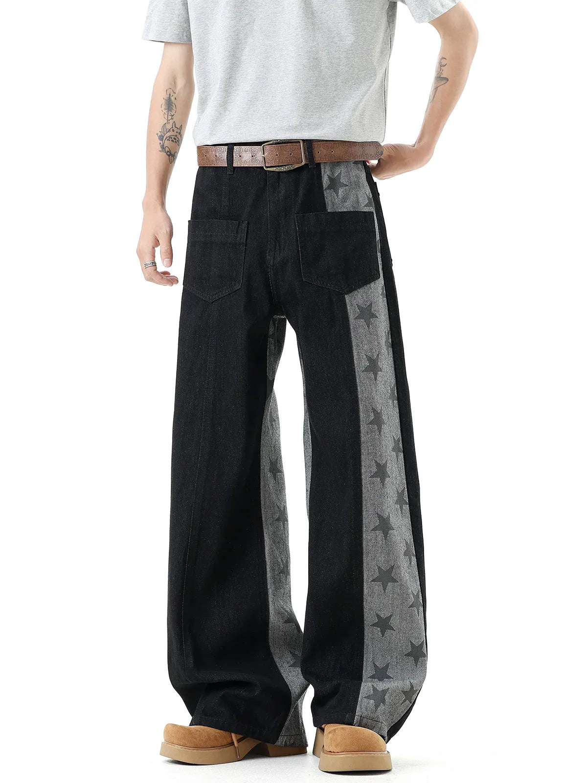 A person wearing wide-leg black denim pants with gray side panels featuring a star pattern, styled with a brown belt, white t-shirt, and tan boots against a white background.