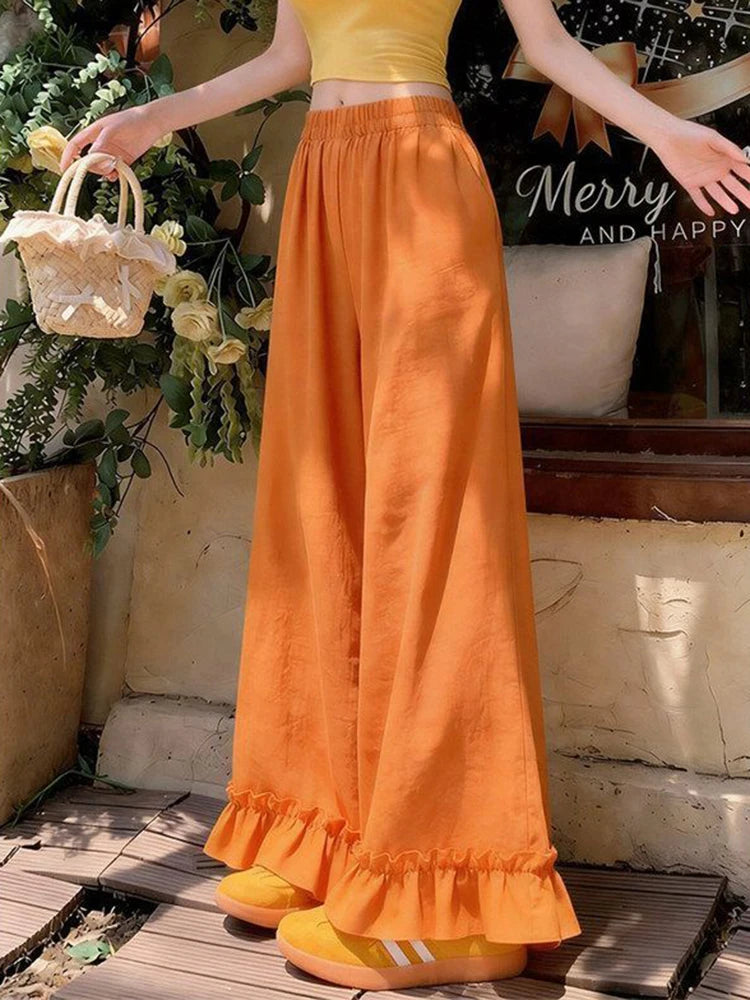 A person wearing bright orange wide-leg ruffled pants and a matching yellow crop top, standing outdoors near a window with flowers and a basket visible.