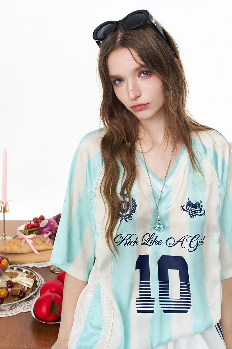 A woman wearing a light blue and white V-neck jersey top with navy graphics, including the text 'Feel Like A Girl' and the number '10'. She has black sunglasses on her head and is posed next to a table with food props against a white studio background.