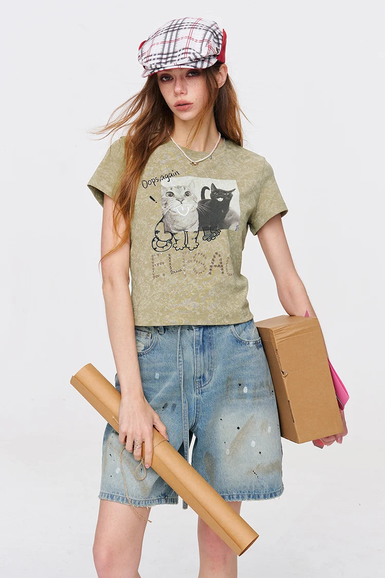 A model wearing a distressed olive green graphic t-shirt with a cat illustration and the text 'DREAMER ELUSA', paired with denim shorts, a plaid flat cap, and holding a cardboard box and a rolled-up paper tube against a plain white background.