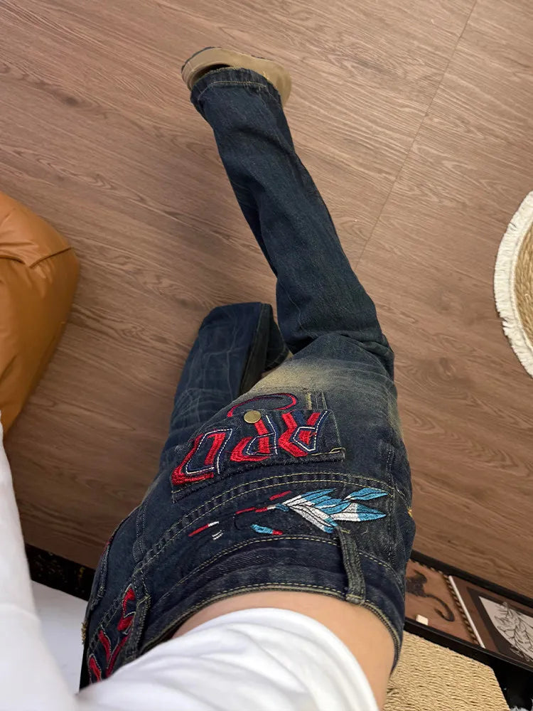 A person wearing dark blue jeans with red and white embroidered lettering and a blue-and-white butterfly patch on the back pocket, standing on a wood-look floor.