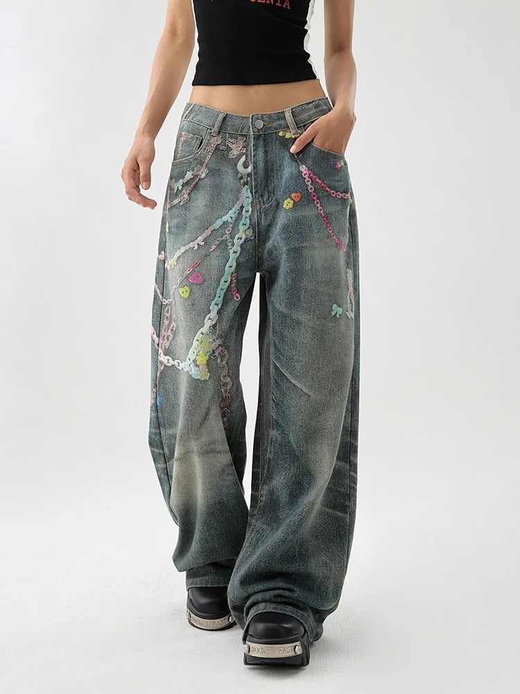 A person wearing very wide-leg distressed denim jeans with colorful chain and bead embellishments, paired with a black cropped top and black platform shoes, photographed against a light gray studio background.