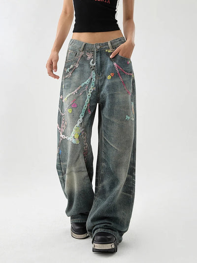 A person wearing very wide-leg distressed denim jeans with colorful chain and bead embellishments, paired with a black cropped top and black platform shoes, photographed against a light gray studio background.