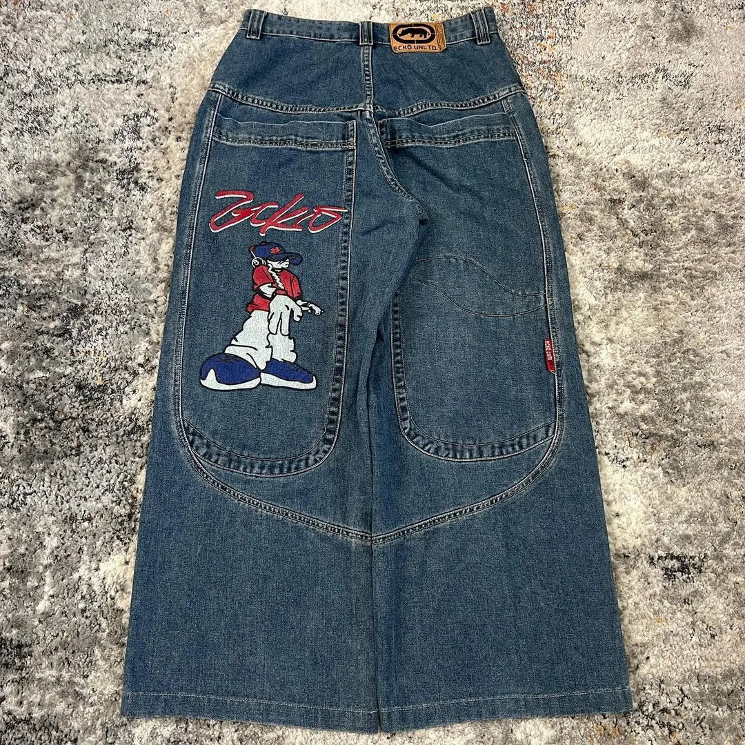 A pair of blue denim jeans laid flat on a textured gray rug, showing the back pockets with a large embroidered cartoon character and red script text on the left pocket.