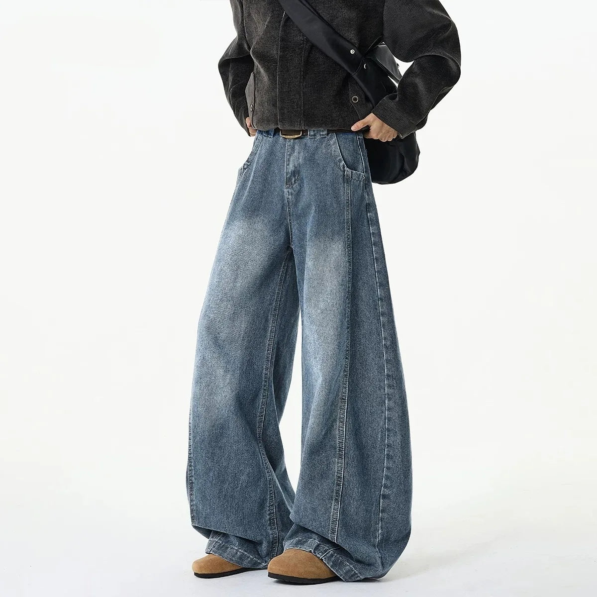 A person wearing very wide-leg light blue denim jeans, styled with a black jacket, black shoulder bag, and brown shoes, photographed against a clean white studio background.