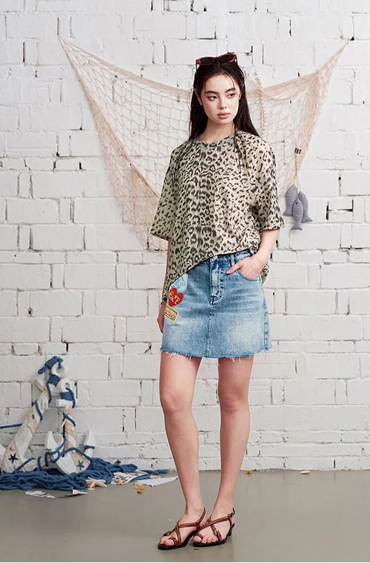 A woman stands against a white brick wall wearing a loose leopard-print top and a light blue denim mini skirt with embroidered details, paired with brown strappy sandals.