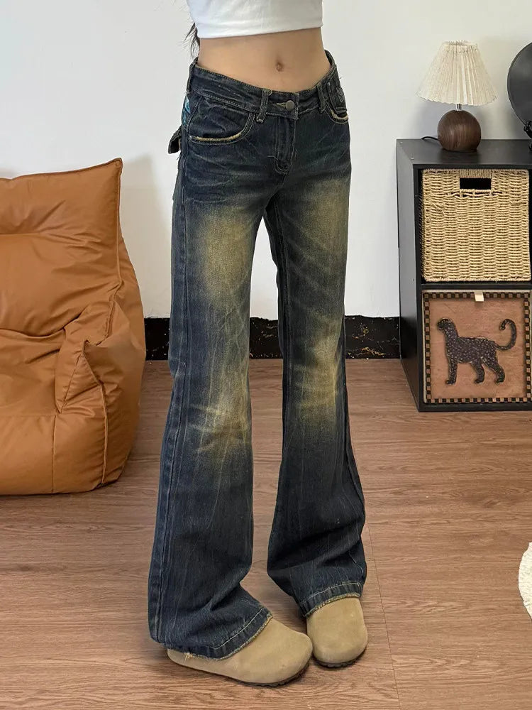 A person wearing dark blue flare jeans with a faded yellow wash, styled with a white crop top and beige slip-on shoes in an indoor room setting.