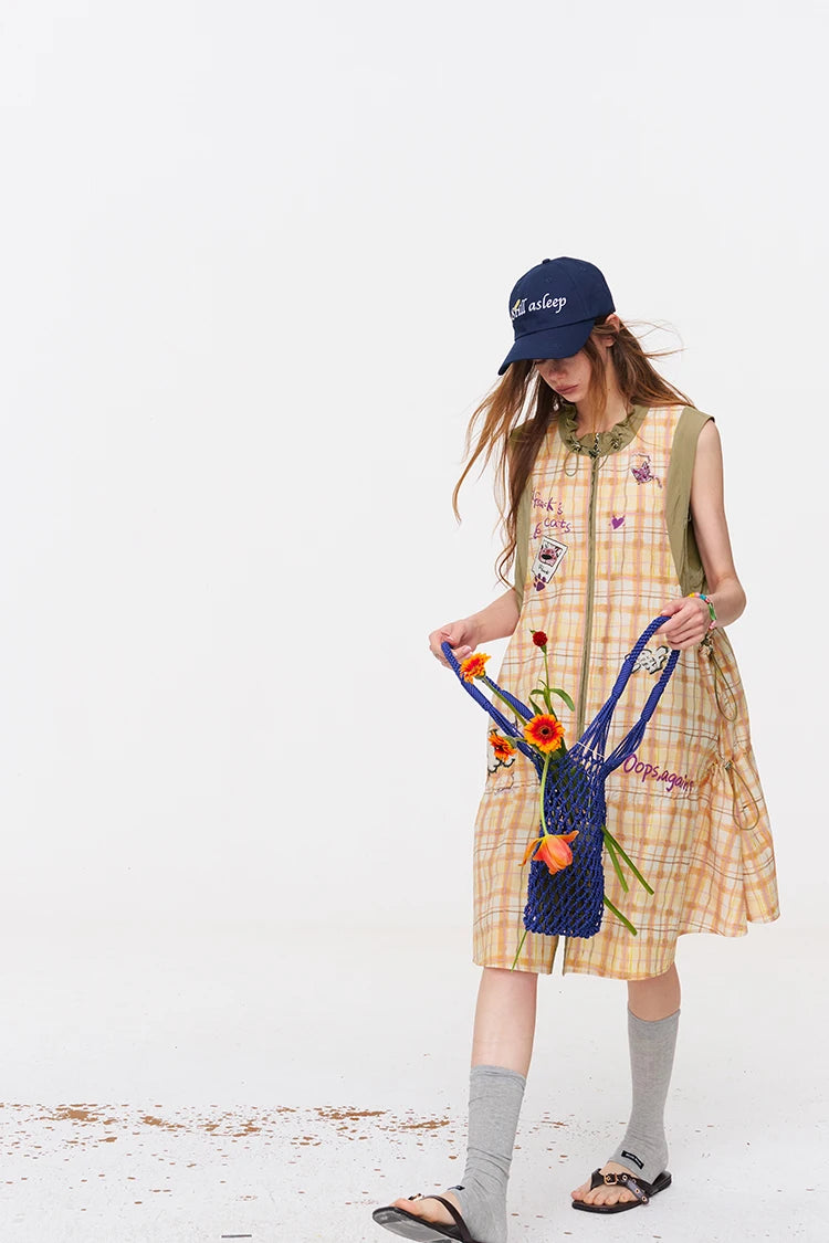 A young woman wearing a yellow and beige plaid sleeveless dress with embroidered details, a navy blue baseball cap, gray knee-high socks, and sandals, holding a blue flower bag with orange tulips against a plain white background.