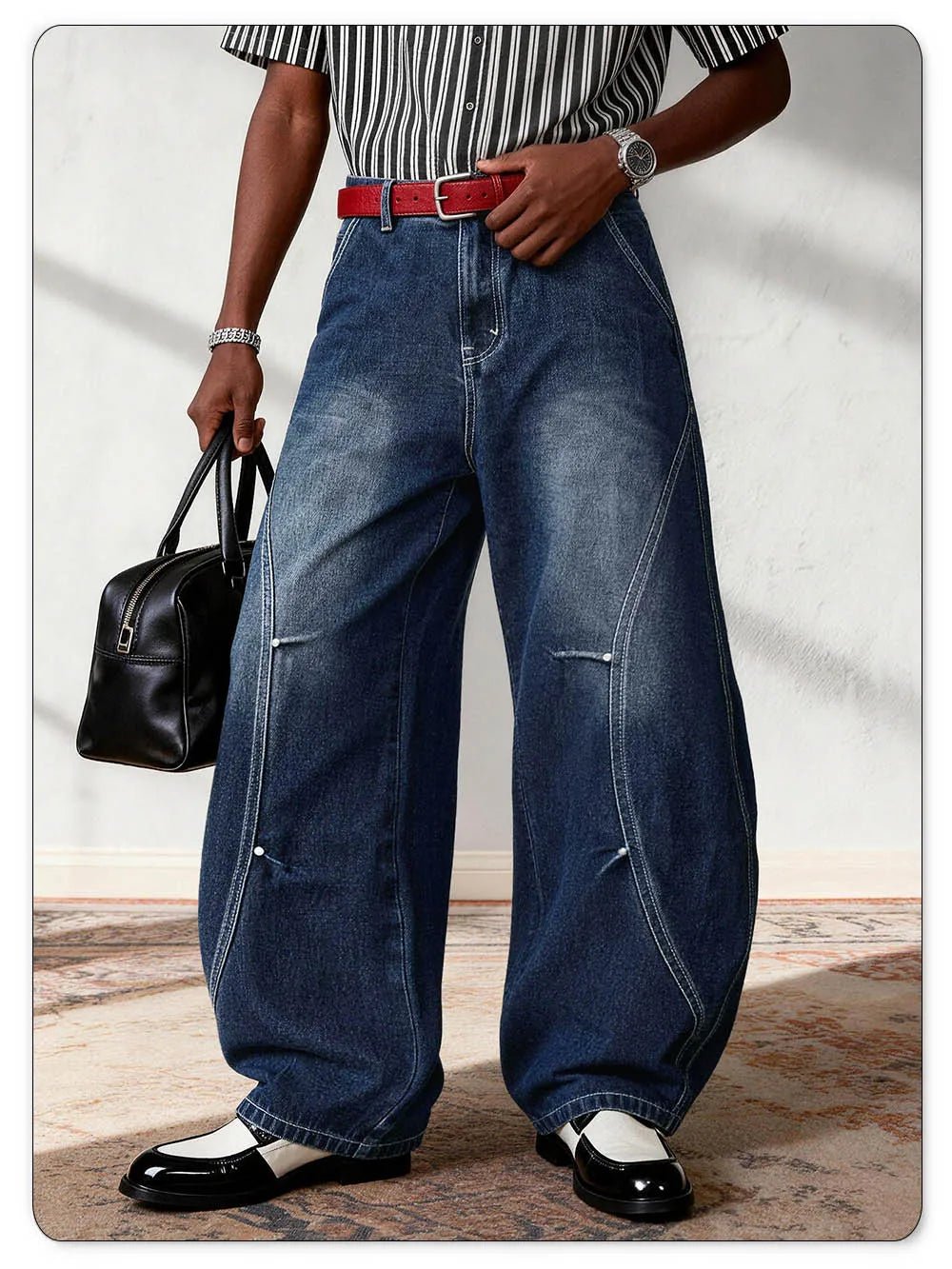 A person wearing oversized dark blue denim jeans with a red belt, a black-and-white striped shirt, and black-and-white shoes, holding a black handbag against a light wall.