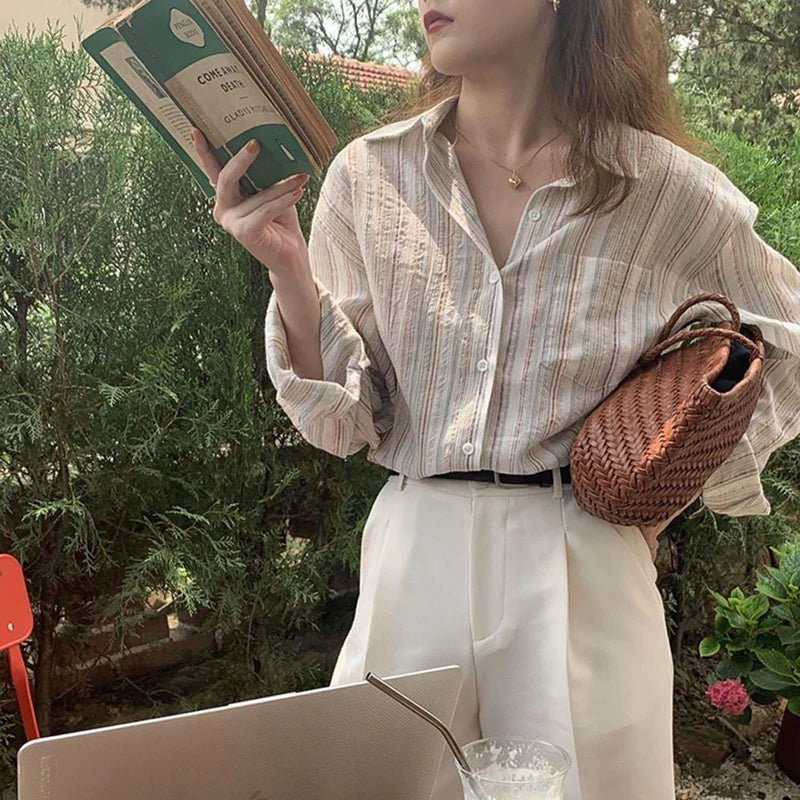 Woman holding a book and a woven bag, sitting outdoors with a laptop and drink.