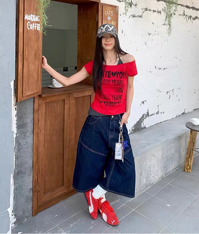 A woman stands in front of a rustic wooden counter at a cafe, wearing a red off-the-shoulder graphic t-shirt, a dark wash wide-leg denim shorts, white socks, and red sneakers.