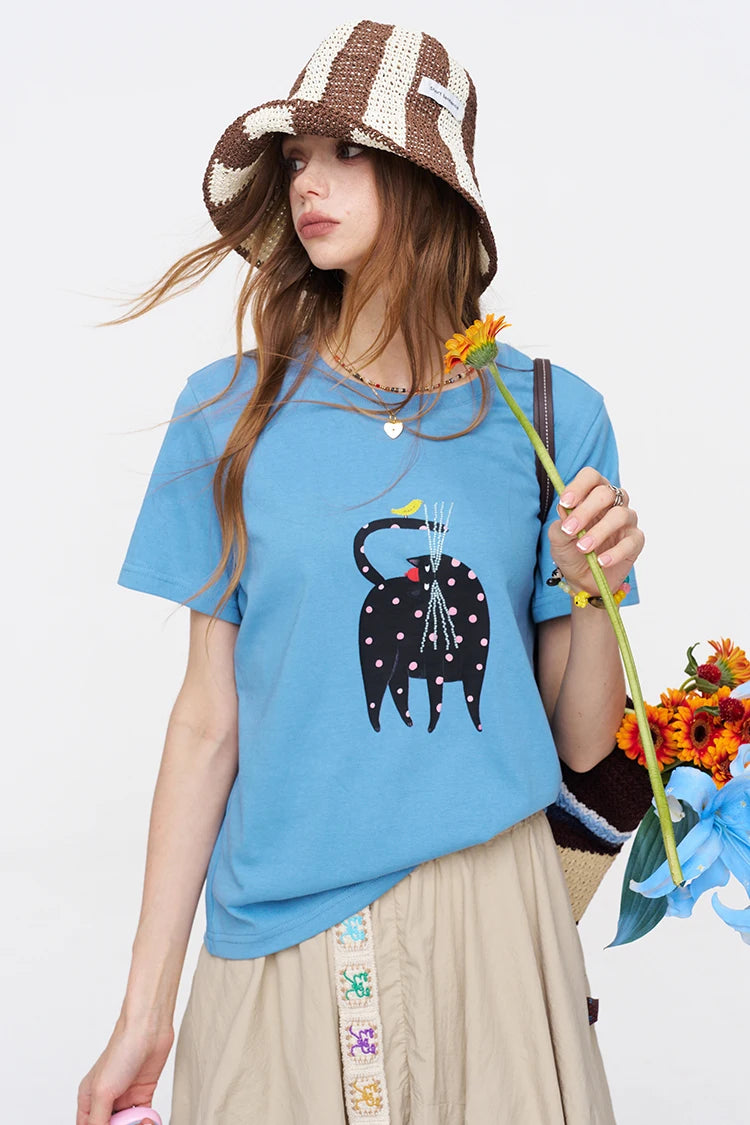 A woman wearing a light blue graphic t-shirt with a black cat design, a brown and beige striped bucket hat, and a beige skirt, holding a yellow flower against a white background.