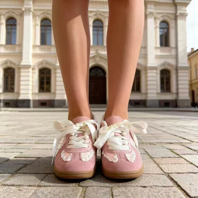 A person wearing pink lace-up sneakers with white bows and white lace appliques, standing on a stone pavement in front of a classical building.