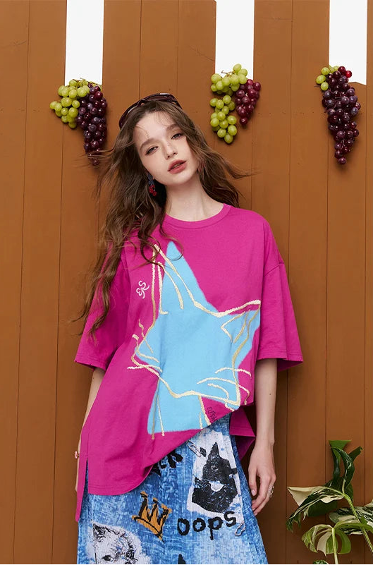 A woman models a bright pink oversized T-shirt with a large light blue star graphic outlined in gold, paired with a blue patterned skirt, set against a brown wooden wall decorated with hanging grape clusters.