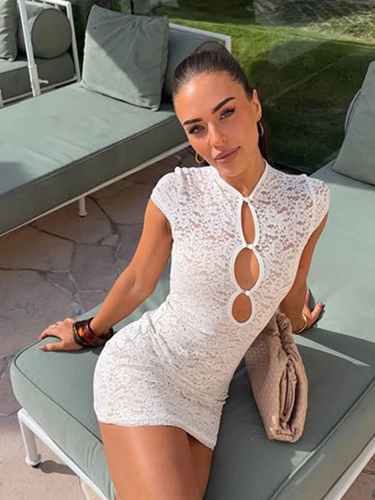 A woman wearing a white lace mini dress with keyhole cutouts sits on a light green outdoor lounge chair next to a beige clutch bag.