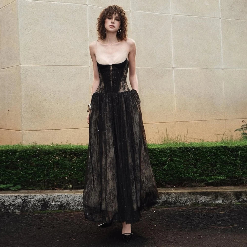 Woman wearing a black lace dress standing against a beige wall with greenery.