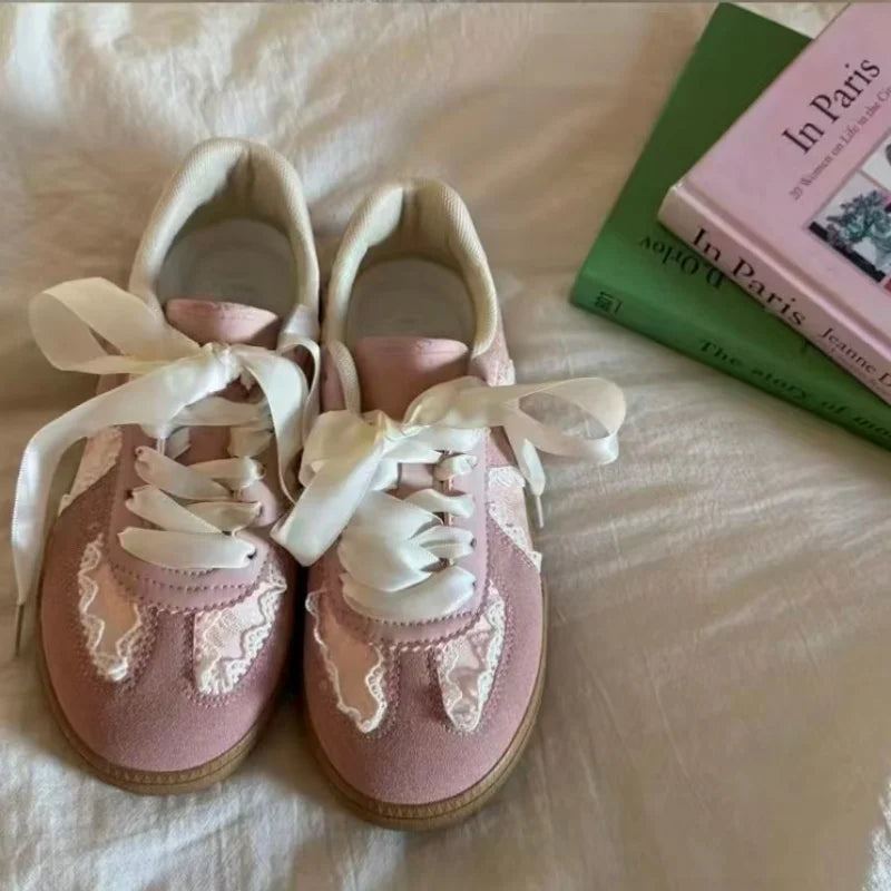 A pair of pink low-top sneakers with white ribbon laces and white lace appliques, resting on a white fabric surface next to a green and pink book.