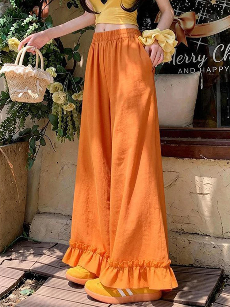 A person wearing bright orange wide-leg pants with a ruffled hem, paired with a yellow crop top and yellow shoes, standing outdoors near a shop window and potted plants.
