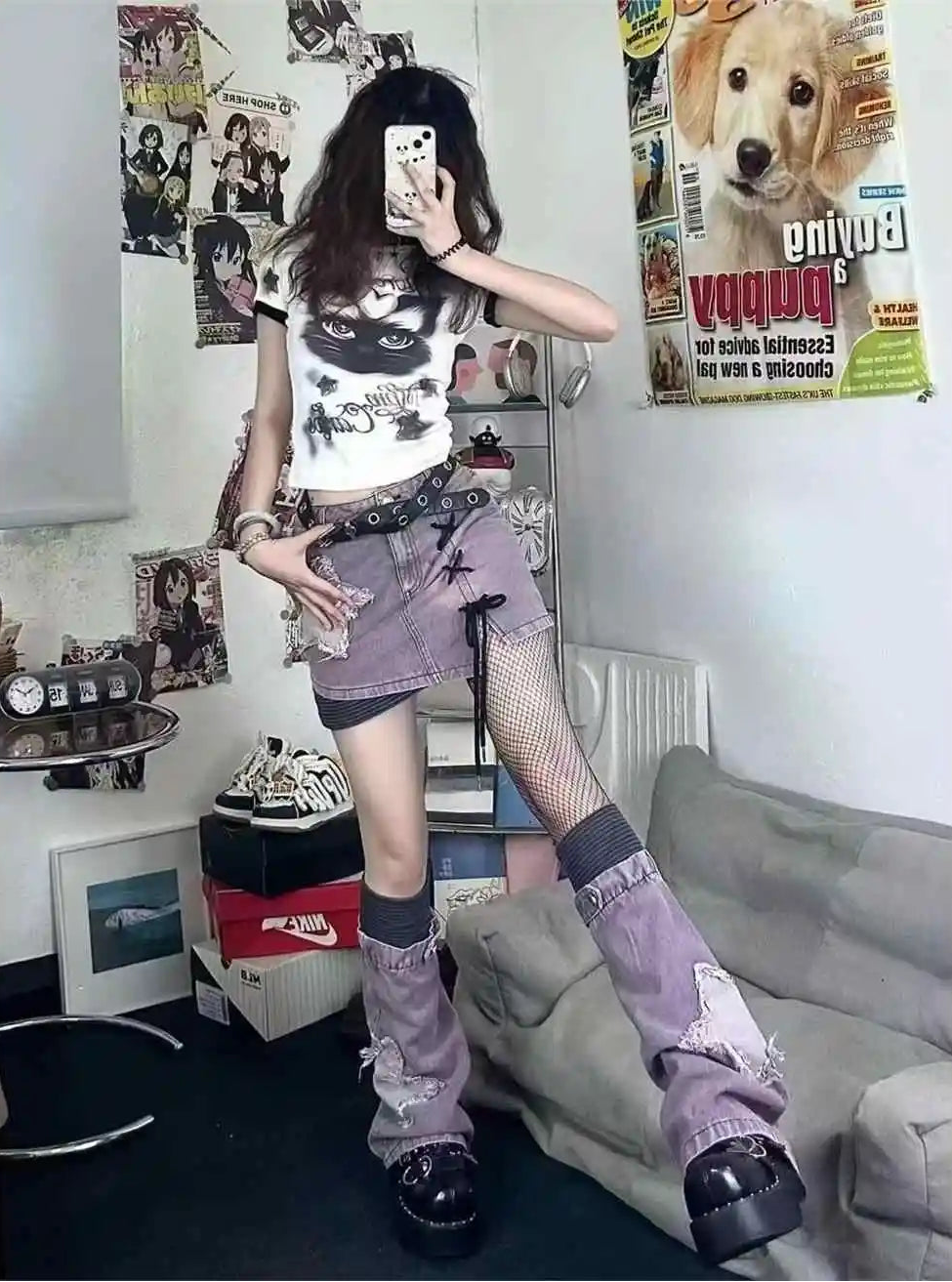 A person takes a mirror selfie in a room, wearing a white graphic t-shirt, a distressed purple denim mini skirt, fishnet stockings, and chunky platform boots. The room contains posters, a gray sofa, and shelves with shoes.