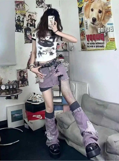A person takes a mirror selfie in a room, wearing a white graphic t-shirt, a distressed purple denim mini skirt, fishnet stockings, and chunky platform boots. The room contains posters, a gray sofa, and shelves with shoes.