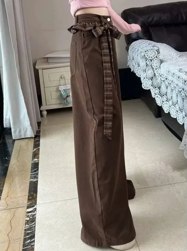 A person wearing high-waisted brown wide-leg pants with a matching brown plaid tie belt, standing in a home interior with a bed and curtain visible in the background.