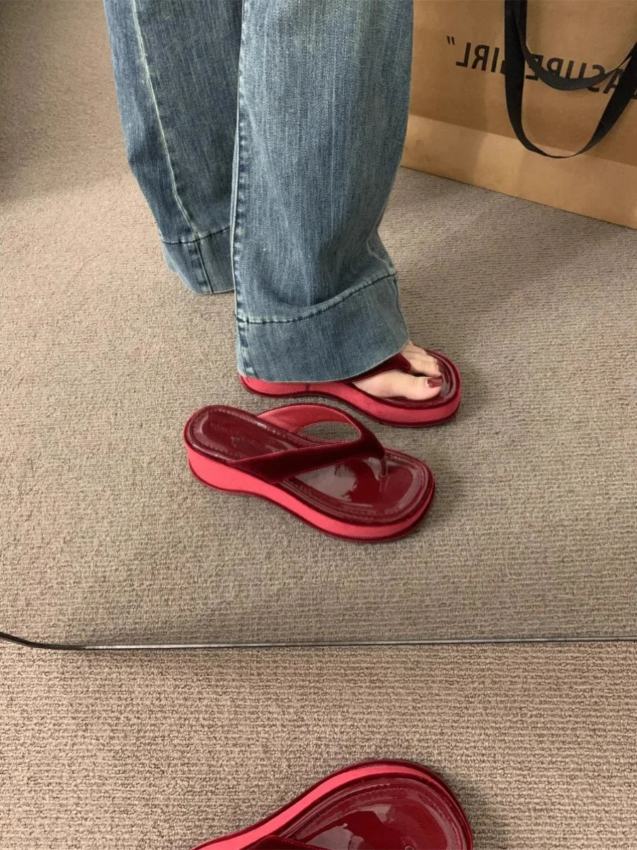 A person wearing translucent red jelly flip-flops and blue jeans, standing on a beige carpet next to a brown paper shopping bag.