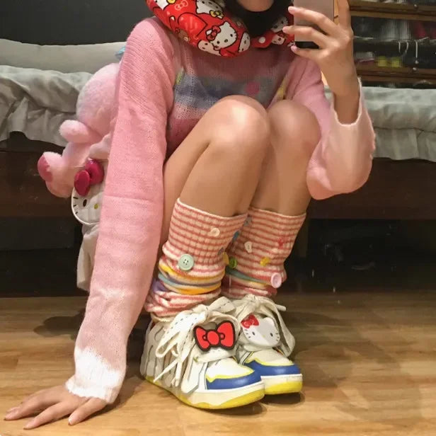 A person crouching in a bedroom setting, wearing a pink fuzzy long-sleeve top, a red Hello Kitty patterned neck warmer, pink and white gingham knee-high socks with colorful stripes, and cream-colored sneakers decorated with Hello Kitty faces and bows.