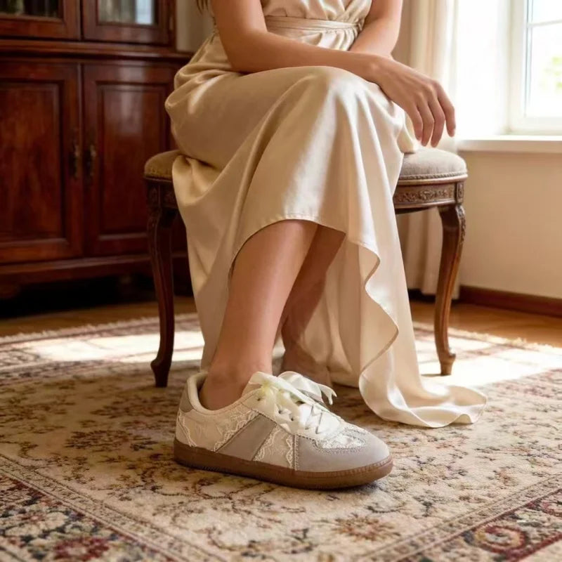 A person seated on a wooden stool in a sunlit room wearing a cream satin dress and light beige low-top sneakers with white lace-up fronts and gray side panels.